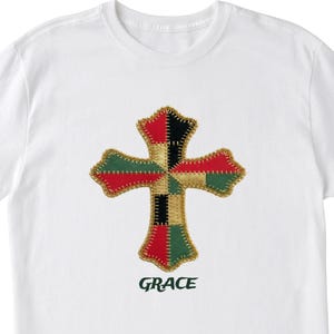 以下が含まれることがあります： カラフルな十字架のデザインが施された白いTシャツ。十字架は赤、緑、黒、金色のステッチパッチでできており、下に緑色の「GRACE」という文字があります。デザインはパッチワークの美学を持っています。