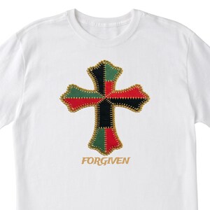 Może przedstawiać: Biała koszulka z krzyżem w patchworku w kolorach czerwonym, czarnym i zielonym, obramowanym złotymi szwami. Słowo "FORGIVEN" jest wydrukowane pod spodem w kolorze złotym. Codzienny element odzieży o tematyce religijnej.