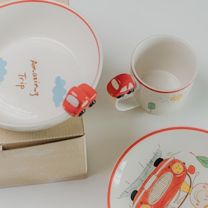 May include: A white ceramic bowl, mug, and plate set with a travel theme. The bowl has the text "Amazing Trip" and a red car accent. The mug has a red car handle and sun and tree illustrations. The plate features a red car illustration.