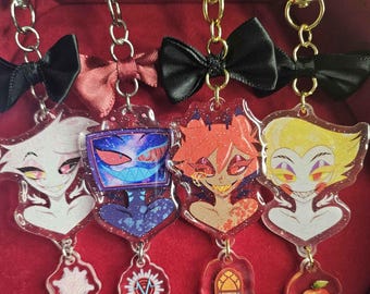 May include: Four acrylic keychains featuring cartoon characters with gold-colored hardware. Each keychain has a different character design, with a small charm at the bottom. The keychains are attached to a red surface.