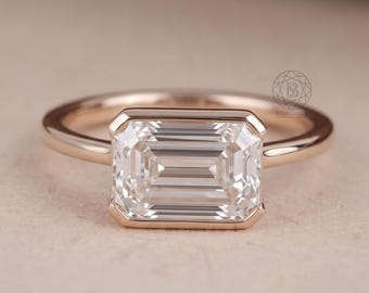 May include: A rose gold ring featuring a rectangular emerald-cut clear gemstone set in a bezel setting. The band is smooth and polished, showcasing a clean, horizontal orientation of the stone. A small circular logo is visible on the band.