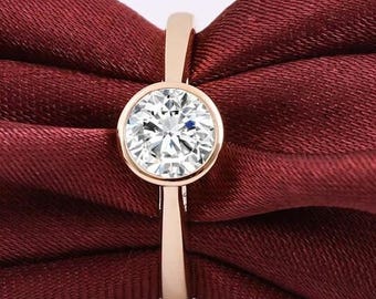 Puede incluir: Un anillo de oro rosa con una gema grande, redonda y transparente engastada en un bisel. El anillo se muestra sobre una tela roja oscura, resaltando el brillo y el diseño del anillo. La banda es delgada y elegante.