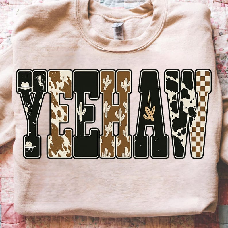 May include: A light pink sweatshirt with the word "YEEHAW" in a bold, decorative font. The letters are filled with various patterns, including cowhide, cacti, and a checkered design. The sweatshirt has a crew neck and long sleeves.