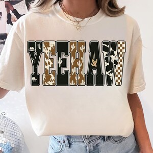 May include: Cream-colored t-shirt with the word "YEEHAW" in a bold, western-style font. The letters feature various patterns, including cow print, cacti, and a checkered design. The shirt has a relaxed fit.