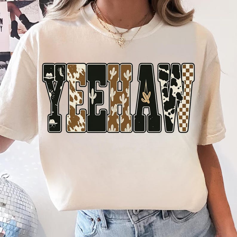 May include: Cream-colored t-shirt with the word "YEEHAW" in a bold, western-style font. The letters feature various patterns, including cow print, cacti, and a checkered design. The shirt has a relaxed fit.