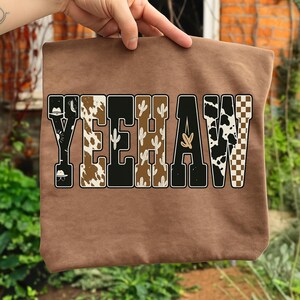May include: A brown tote bag with the word "YEEHAW" in a Western-themed design. The letters are filled with patterns like cowhide, cacti, and a checkered pattern. The bag is held up by a hand.