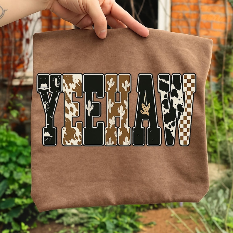 May include: A brown tote bag with the word "YEEHAW" in a Western-themed design. The letters are filled with patterns like cowhide, cacti, and a checkered pattern. The bag is held up by a hand.