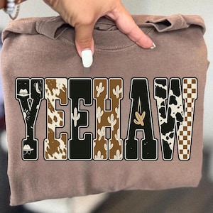 May include: A brown sweatshirt with the word "YEEHAW" in a bold, western-style font. The letters feature various patterns, including cowhide, cacti, and a checkered design, with cowboy hat illustrations.
