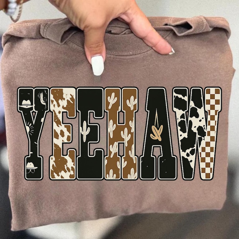 May include: A brown sweatshirt with the word "YEEHAW" in a bold, western-style font. The letters feature various patterns, including cowhide, cacti, and a checkered design, with cowboy hat illustrations.