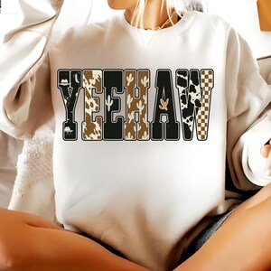 May include: A white sweatshirt with the word "YEEHAW" in a western-themed design. The letters are decorated with a cowboy hat, cow print, cacti, and a checkered pattern. The sweatshirt is worn by a person.