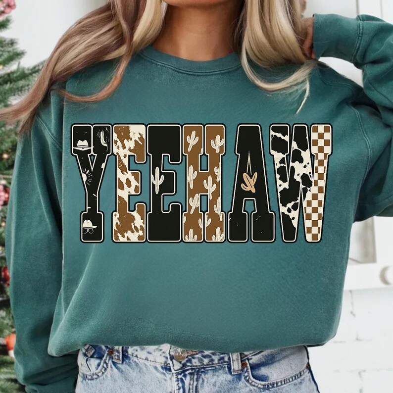 May include: Teal sweatshirt with the word "YEEHAW" in a western-themed design. The letters feature patterns like cow print, cacti, and a checkered pattern. The sweatshirt is a solid teal color.
