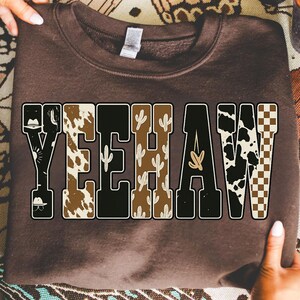 May include: A brown sweatshirt with the word "YEEHAW" in a bold, block letter design. The letters are filled with various patterns, including cowhide, cacti, and a checkered pattern. The sweatshirt is held up by two hands.