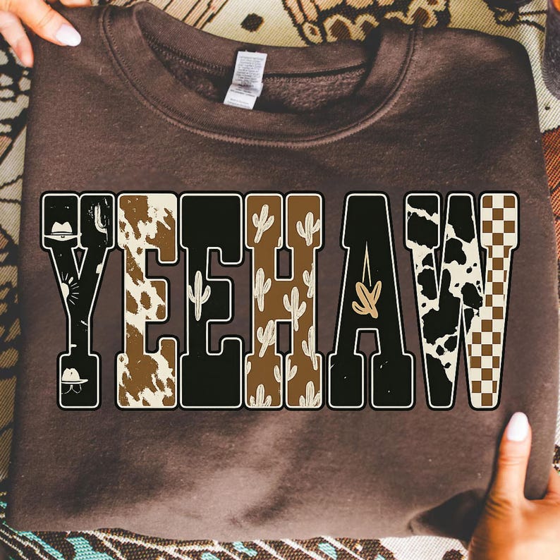 May include: A brown sweatshirt with the word "YEEHAW" in a bold, block letter design. The letters are filled with various patterns, including cowhide, cacti, and a checkered pattern. The sweatshirt is held up by two hands.