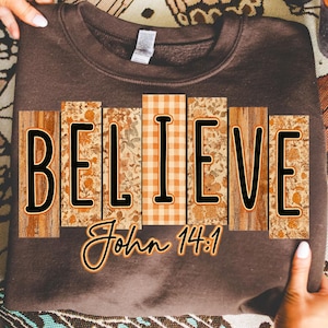 Könnte beinhalten: Ein braunes Sweatshirt mit dem Wort "BELIEVE" in einem Blockbuchstaben-Design. Die Buchstaben bestehen aus Holz-, Blumen- und Karomustern. Der Text "John 14:1" steht darunter.