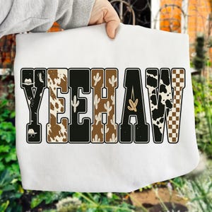 May include: White sweatshirt featuring the word "YEEHAW" in a Western-themed design. The letters are in various patterns, including cow print, cactus, and a checkered pattern. The design includes a cowboy hat.
