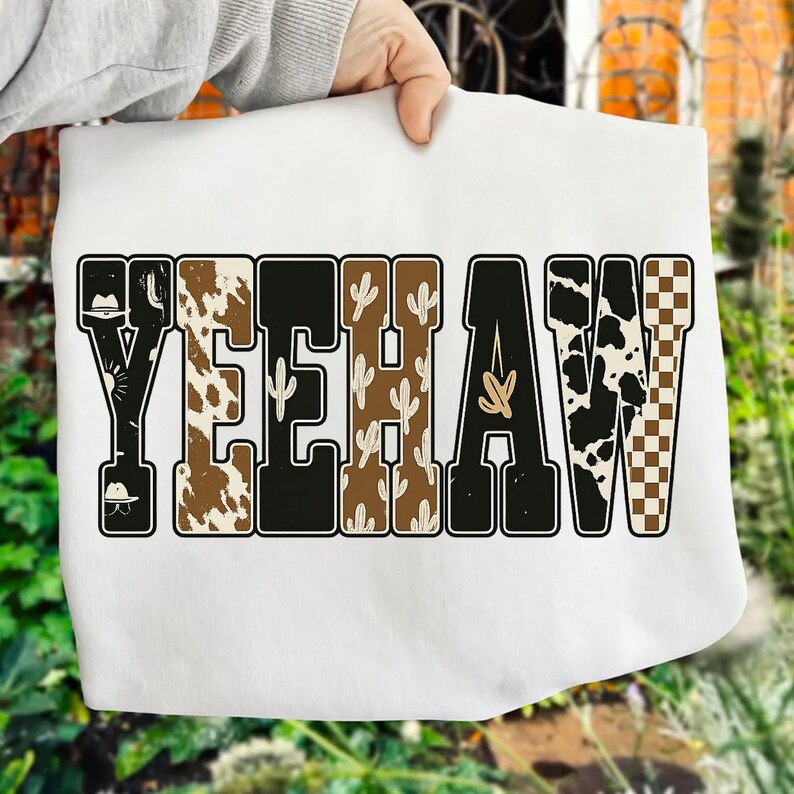 May include: White sweatshirt featuring the word "YEEHAW" in a Western-themed design. The letters are in various patterns, including cow print, cactus, and a checkered pattern. The design includes a cowboy hat.