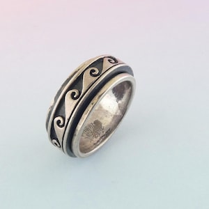 May include: A silver-colored ring with a wave pattern design. The ring has a spinning outer band with a black background and a repeating wave pattern. The ring is made of metal and has a polished finish.