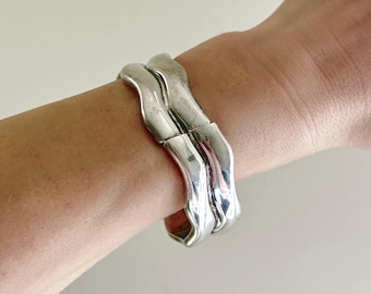 May include: A silver-colored cuff bracelet with a wavy design. The bracelet is worn on a wrist and has a polished, reflective surface. The bracelet appears to be made of metal and has a modern, sculptural aesthetic.