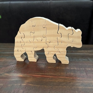 May include: A wooden bear puzzle with interlocking pieces. The bear is light-colored, with visible wood grain. The puzzle is assembled and stands on a dark wooden surface. The bear has a simple, stylized design.