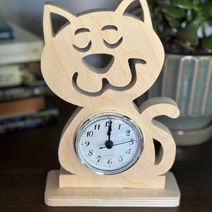 May include: A wooden cat-shaped clock with a round clock face. The clock is made of light-coloured wood and features a smiling cat face with closed eyes. The clock face has black numbers and hands. The clock is a decorative item.