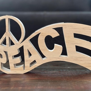 Op de afbeelding: Een houten vredesteken met het woord "PEACE" uitgesneden. Het vredessymbool staat links en het woord staat in een gebogen banier. Het hout heeft een natuurlijke, lichtbruine kleur.