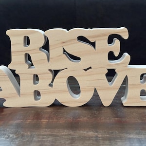 Könnte beinhalten: Holzschild mit den Worten "RISE ABOVE" in einer dekorativen Schriftart ausgeschnitten. Das Schild besteht aus hellfarbenem Holz und steht auf einer dunklen Holzoberfläche. Die Buchstaben sind verbunden und haben einen 3D-Effekt.