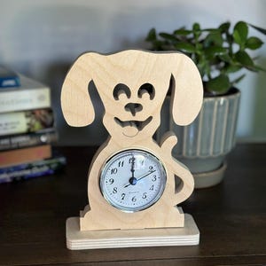 Puede incluir: Un reloj de madera con forma de perro y esfera redonda. El reloj es de madera clara y tiene un diseño de cara de perro sonriente. La esfera tiene números y manecillas negras. El reloj está sobre una base de madera.