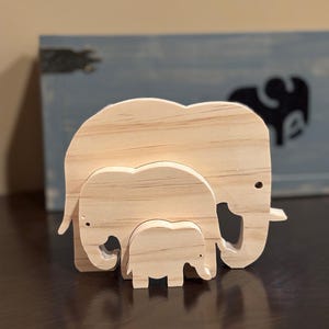 May include: A wooden elephant family sculpture. The set includes a large, medium, and small elephant, all crafted from light-colored wood. The elephants are arranged together, showcasing a family theme. The sculpture is a decorative item.
