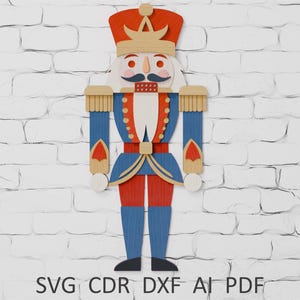 以下が含まれることがあります： 赤、青、ベージュの木製クリスマスくるみ割り人形。くるみ割り人形は、金の王冠が付いた赤い帽子、金のトリムが付いた青いジャケット、赤いズボンを着用しています。画像には「SVG CDR DXF AI PDF」というテキストが含まれています。