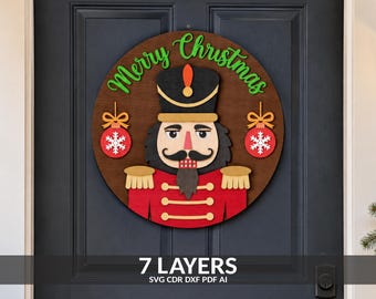 May include: A circular wooden door sign featuring a layered nutcracker design in red, black, and tan. The text Merry Christmas is written in green script at the top. Two red ornaments with white snowflakes hang on either side of the nutcracker figure.