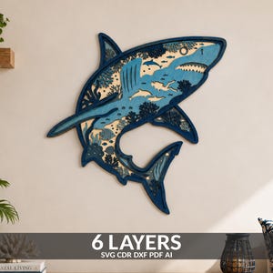 Op de afbeelding: Een meerlaags houten wandkunstwerk in de vorm van een haai, met ingewikkelde uitsparingen van koraalriffen en kleinere vissen. Het ontwerp maakt gebruik van verschillende blauwtinten en natuurlijke houttinten, wat een ruimtelijk effect creëert met zes lagen.