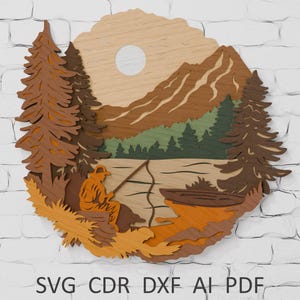 May include: Wooden layered art piece depicting a serene fishing scene. The artwork features a fisherman, mountains, trees, and a lake, all in shades of brown, green, and orange. A sun is visible in the background. The text "SVG CDR DXF AI PDF" is at the bottom.