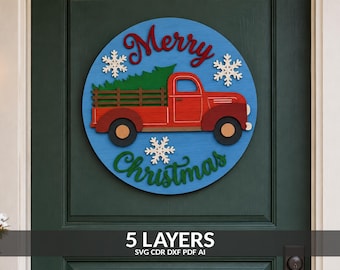 May include: A circular wooden sign featuring a blue background with a red truck carrying a green Christmas tree. The text Merry is in red script at the top and Christmas is in green script at the bottom, accented by white snowflakes.