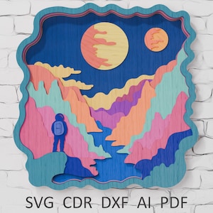 May include: A layered wooden art piece depicting a colorful space scene. The artwork features an astronaut, mountains, two planets, and a blue sky. The piece is framed with a wavy, teal border. The bottom of the image includes the text "SVG CDR DXF AI PDF".