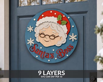 May include: A circular wooden door sign featuring a layered illustration of Mrs. Claus wearing a red hat with white trim and holly. The blue background includes white snowflakes and the red text Santa's Boss. The design consists of nine layers.