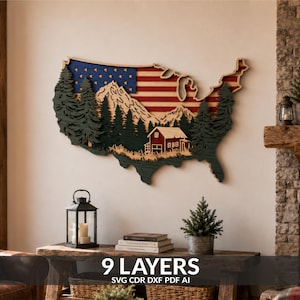 May include: A multi-layered wooden wall art piece shaped like the map of the United States. The design features a red, white, and blue flag pattern, a mountain range, a forest of dark green pine trees, and a small red cabin in the foreground.