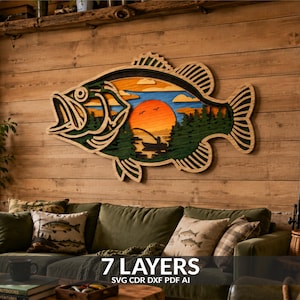 Peut inclure: Une décoration murale en bois multicouche en forme de grand poisson, présentant un motif découpé au laser d'un coucher de soleil sur un lac avec la silhouette d'une personne pêchant en bateau, entourée de pins, dans des tons orange, bleu et vert.