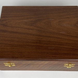 Puede incluir: Una caja rectangular de madera de color marrón oscuro, con un patrón de vetas naturales. La caja está asegurada con dos cierres dorados. La tapa está cerrada, lo que sugiere que el contenido de la caja está oculto.