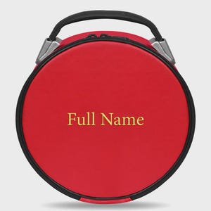 Op de afbeelding: Een ronde, rode koffer met een zwarte rits en handvat. De tekst "Full Name" is in goud op de voorkant geborduurd. De handgreep is zwart en de hardware is zilver.