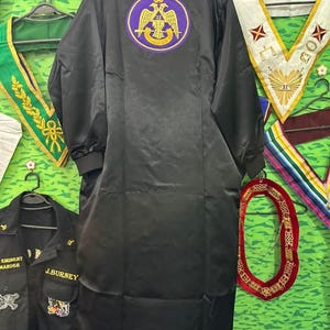 Puede incluir: Bata de satén negro con cuello morado y un emblema circular en la espalda. El emblema presenta un águila dorada y otros símbolos dentro de un círculo morado. Se ven otros artículos ceremoniales, como una faja verde y una camisa negra.