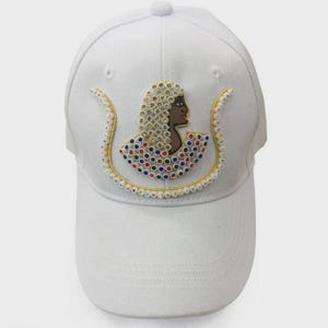 Könnte beinhalten: Weiße Baseballkappe mit einem gestickten Design eines Profils einer Person mit Perlenkopfschmuck und Kragen. Das Design ist mit farbigen Strasssteinen und goldfarbenen Zierleisten verziert.