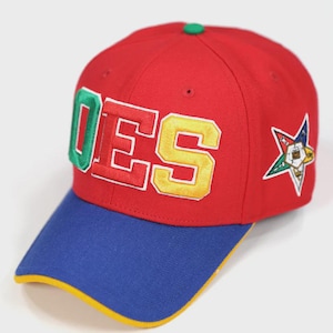 Puede incluir: Gorra de béisbol roja con visera azul y ribete amarillo. La gorra presenta la palabra "OES" en letras de bloque verdes, amarillas y rojas. Un emblema de estrella está en el lateral. Botón verde en la parte superior.