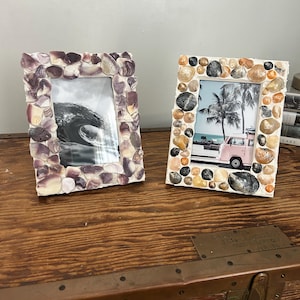 May include: Two rectangular picture frames decorated with natural shells. The left frame features purple and white shells surrounding a black and white ocean wave photo. The right frame has tan, brown, and grey shells around a beach scene with a pink van.
