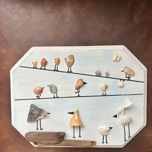 May include: A handcrafted wall art piece featuring seashell birds perched on black lines against a light blue background. The birds are made from various shells and are mounted on a white, octagonal wooden board. A driftwood piece is at the bottom.