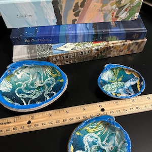 May include: Hand-painted seashell art featuring octopus and turtle designs in blue and white. The shells have blue outlines and are displayed on a black surface. Books with titles such as "Jane Eyre" and "The Great Gatsby" are stacked in the background.