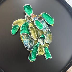 Puede incluir: Una escultura decorativa de tortuga con acabado metálico. La tortuga presenta una mezcla de tonos verdes, amarillos y azules, con un contorno plateado. La escultura está colocada sobre una bandeja redonda negra, creando un contraste llamativo.