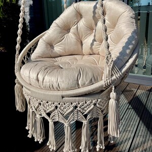 Peut inclure: Un fauteuil suspendu en macramé beige avec un coussin moelleux et capitonné. La chaise présente un design tissé avec des glands décoratifs et est suspendue par d'épaisses cordes tressées. La chaise est conçue pour la détente.