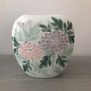 May include: A white ceramic vase with a rounded shape, adorned with hand-painted floral designs. The flowers are in shades of pink and lavender, with green leaves. The vase is a decorative item.