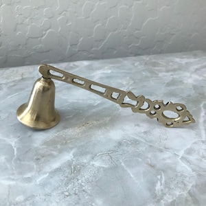 May include: A brass bell with a decorative handle. The bell is a classic shape, with a flared base and a rounded top. The handle is ornate, with cut-out designs. The bell is resting on a marble-patterned surface.