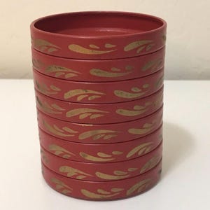 May include: A stack of eight red coasters with a gold leaf design. The coasters are cylindrical and have a slightly raised edge. The gold leaf design is repeated around the edge of each coaster, creating a decorative pattern.